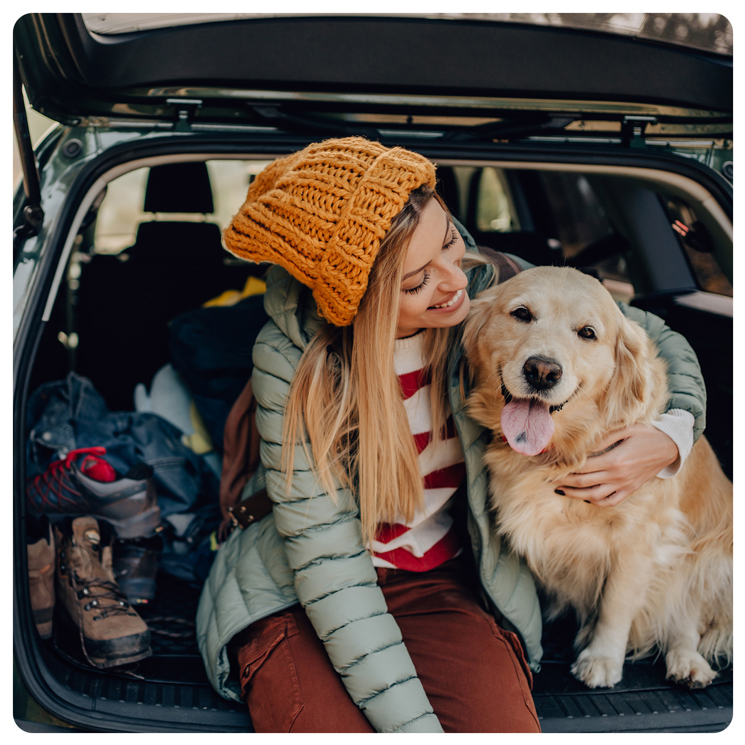 Eine Frau mit gelber Strickmütze und grüner Jacke umarmt einen glücklichen Golden Retriever, während sie im offenen Kofferraum eines Autos sitzt, das mit Outdoor-Ausrüstung, Reiseutensilien und Wanderschuhen beladen ist.