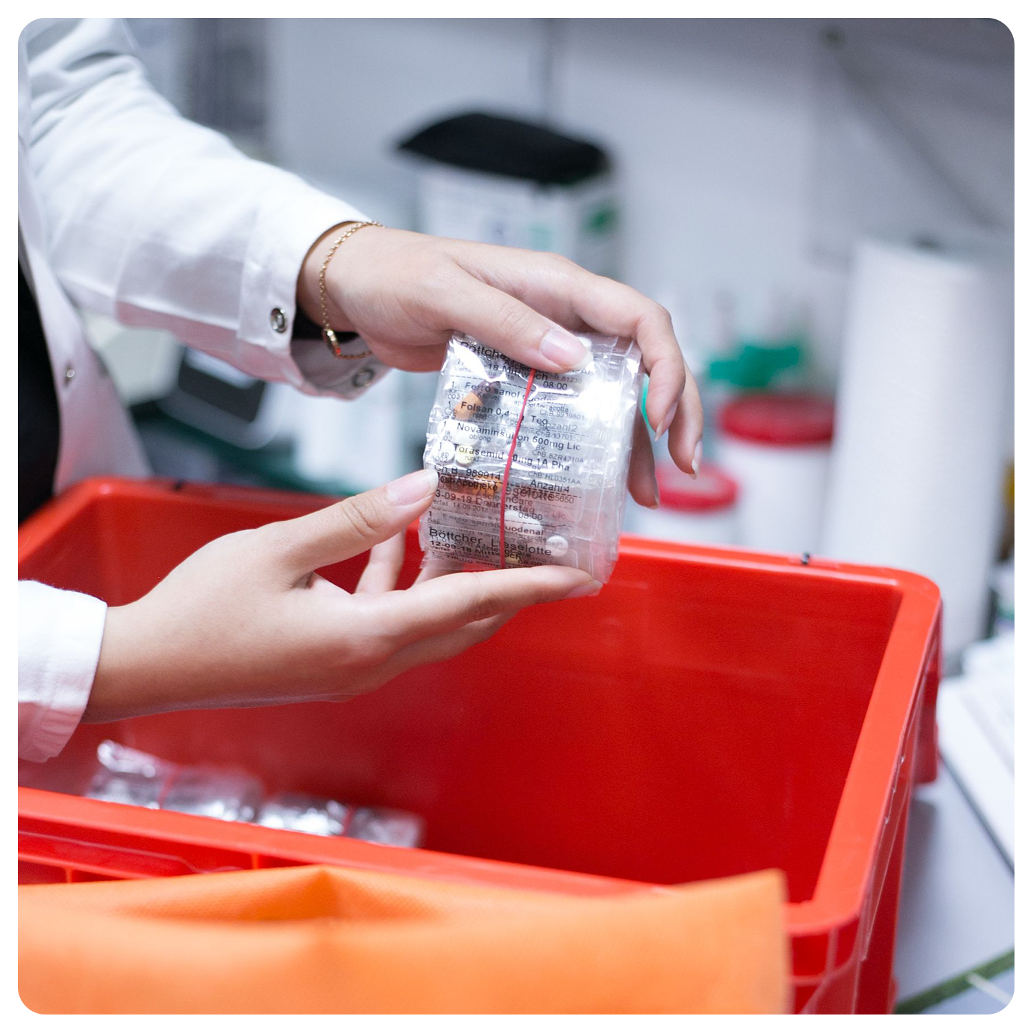 Eine Person im Laborkittel legt verpacktes medizinisches Material, das von der Verblisterung Kassel frisch zubereitet wurde, in einen roten Biohazard-Behälter auf einen überfüllten Labortisch.