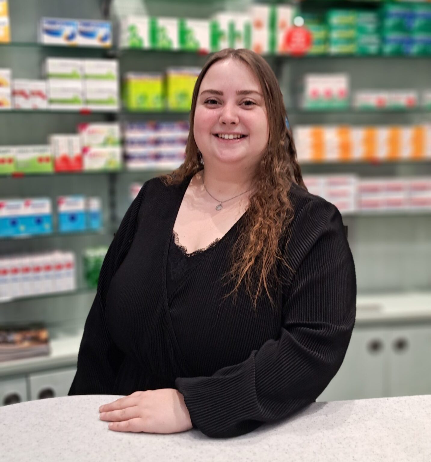 Eine lächelnde Frau mit langen braunen Haaren, die ein schwarzes Oberteil trägt, steht hinter einem Tresen in einer Apotheke, möglicherweise während ihrer Ausbildung in der Apotheke, mit Regalen mit verschiedenen Medikamentenpackungen im Hintergrund.