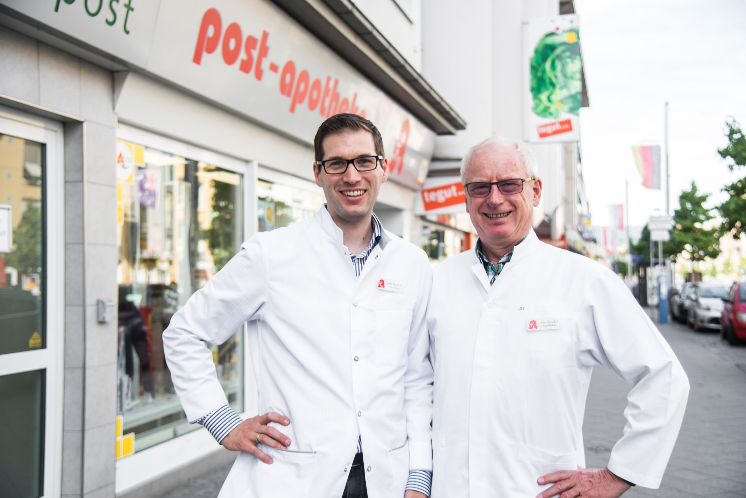 Zwei männliche Apotheker in weißen Laborkitteln stehen lächelnd vor einem Apothekenladen mit der Aufschrift post-apotheke in einer Stadtstraße und betonen damit ihre Verbindung zum Sterillabor Kassel.